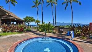 A swimming pool with a tiled edge, surrounded by palm trees and a building with a tiled roof. - Remodeled 2BD Condo w AC Pool Hot Tub-Papakea H106 (Lahaina)