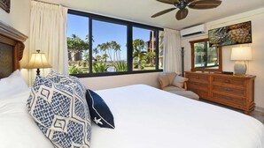 A hotel room with a large bed, a ceiling fan, a dresser, and a view of palm trees and a beach. - Remodeled 2BD Condo w AC Pool Hot Tub-Papakea H106 (Lahaina)