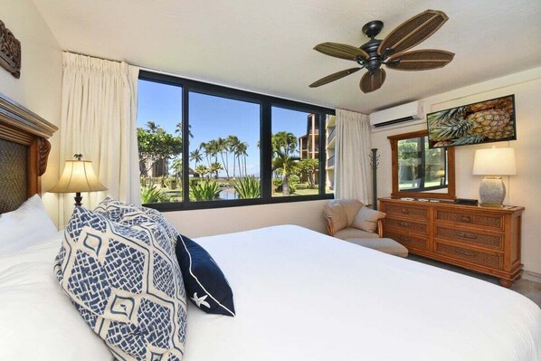 Habitación de hotel con una cama grande, ventilador de techo, cómoda y vista a palmeras y una playa.