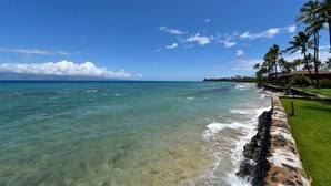 A beachfront with clear turquoise waters, a stone wall, and palm trees. - Remodeled 2BD Condo w AC Pool Hot Tub-Papakea H106 (Lahaina)