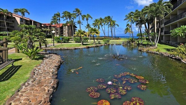 A hotel courtyard with a pond, palm trees, and a view of the ocean. - Remodeled 2BD Condo w AC Pool Hot Tub-Papakea H106 (Lahaina)