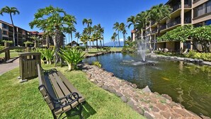 A hotel courtyard with a pond, palm trees, a bench, and a building with balconies. - Remodeled 2BD Condo w AC Pool Hot Tub-Papakea H106 (Lahaina)