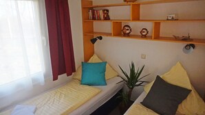 Une petite pièce agréable et chaleureuse, avec un lit, une étagère remplie de livres et d’objets décoratifs, une fenêtre avec des rideaux et une plante.