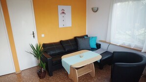 Un salon avec un canapé noir, une table basse en bois, une plante en pot, un tableau encadré au mur et une fenêtre ornée de rideaux légers.