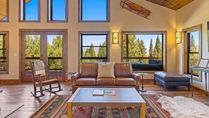 Amplia sala de estar con un sofá de cuero, un sillón mecedora y una mesa de centro. Amplias ventanas ofrecen vistas a árboles y un balcón.