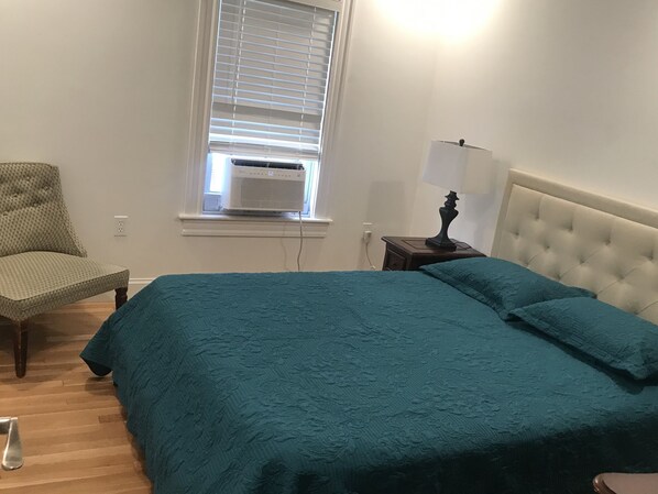 A bedroom with a bed, a chair, a nightstand, and a window with an air conditioner. - Tranquil 1875 Victorian with beautiful grounds and inground pool (South Kingstown)