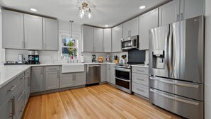 A modern kitchen with grey cabinets, stainless steel appliances, a wooden floor, and a window with a view of greenery. - Tranquil 1875 Victorian with beautiful grounds and inground pool (South Kingstown)