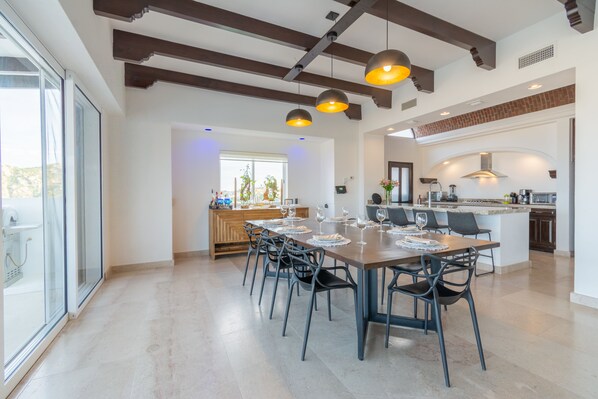 A spacious dining area with a long table and chairs, a kitchen in the background, and a large window providing natural light.