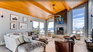 A spacious living room with a fireplace, large windows, and a high ceiling with wooden beams. - Colorado's Coolest Ski In/Out Home - World Class Mtn. Biking, Hiking & Fishing (Granby)