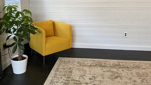 Een kamer met een geel fauteuil, een grote potplant, een wit boekenrek en een hangplant.