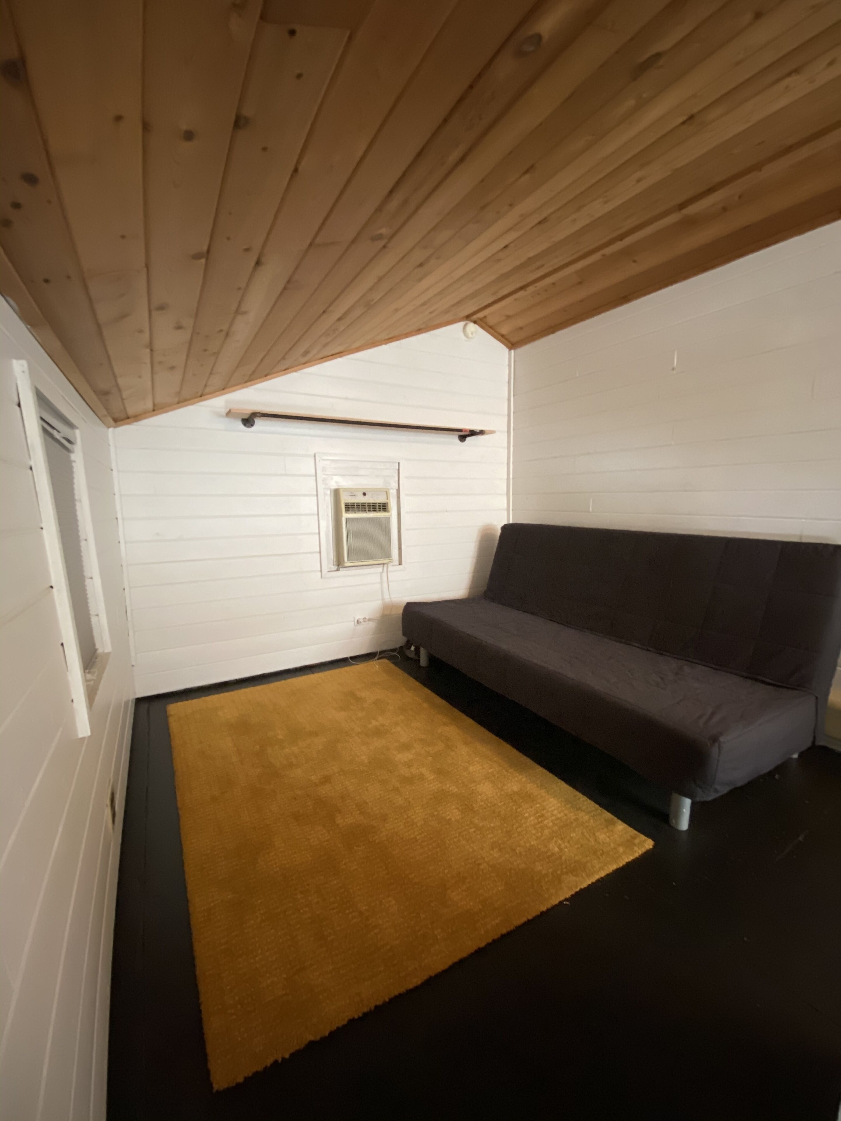 A room with a dark brown floor, a brown rug, a dark brown sofa, a white wall with a window, and a ceiling with wooden beams.
