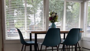 Een eetruimte met een tafel en vier stoelen, grote ramen met jaloezieën en een vaas met bloemen op de tafel.