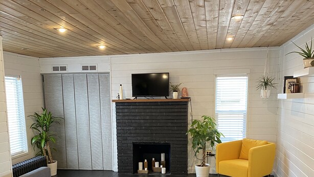 Een moderne woonkamer met een grijze bank, een zwarte open haard, een gele fauteuil en een tv op een houten schoorsteenmantel.