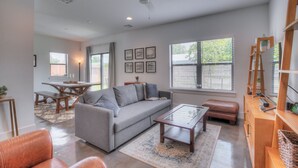 Ein geräumiges Wohnzimmer mit grauem Sofa, Couchtisch und Pouf, ein Essbereich mit Holztisch und Bänken sowie ein Bücherregal mit diversen Gegenständen.