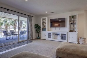 A living room with a large flat-screen TV, white cabinets, a sliding glass door leading to an outdoor patio with chairs and a table, and a potted plant.