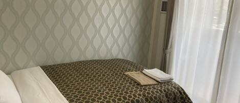 A hotel room with a patterned bedspread, a neatly made bed with a folded towel, and a window with sheer curtains.