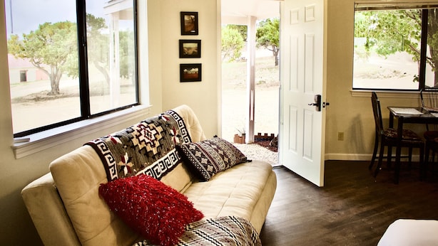 A living room with a beige sofa, a red throw pillow, and patterned cushions. There are three framed pictures on the wall, a dining table with chairs, and a door leading outside. - Authentic Native American Hogan Experience near Chaco Cultural Historical Park (Counselor)