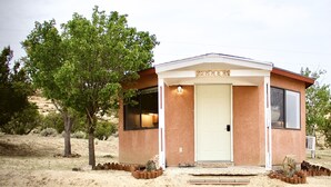 A small, single-story building with a beige exterior and a white door. It has a small porch with a light fixture above the door and a window on the front wall. - Authentic Native American Hogan Experience near Chaco Cultural Historical Park (Counselor)