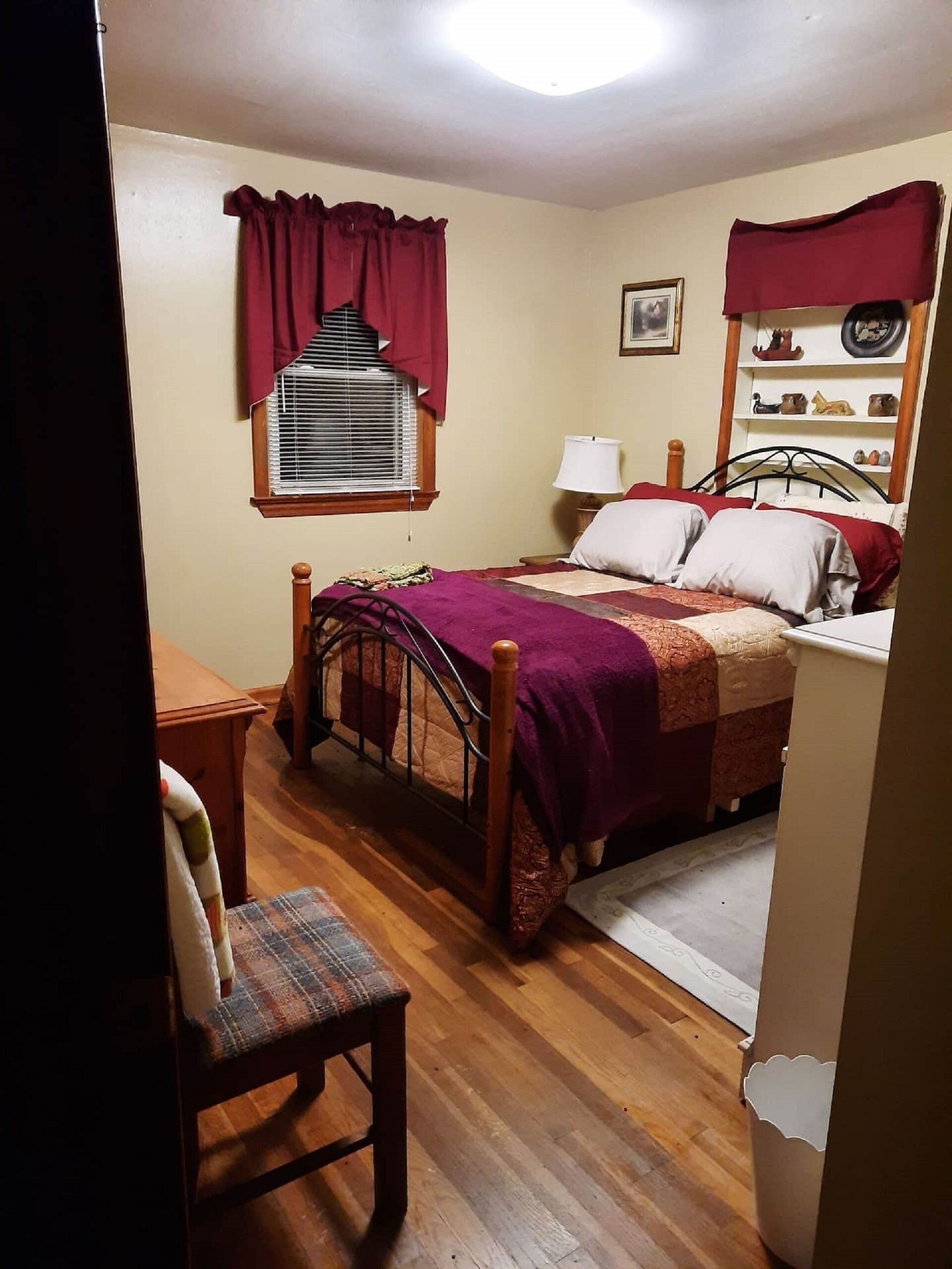Un dormitorio con una cama de madera, una silla, una ventana con cortinas, una mesita de noche y una lámpara.