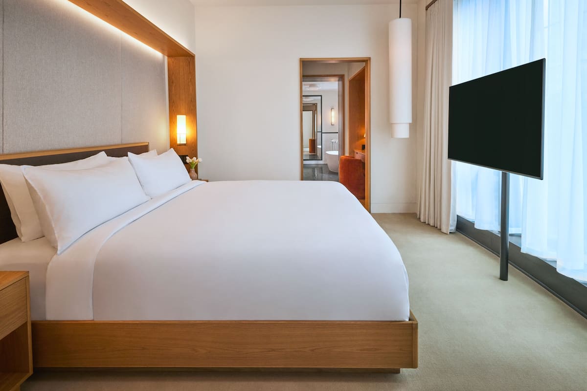 A modern hotel room with a large bed, a flat-screen TV, and a view of the bathroom through an open door.