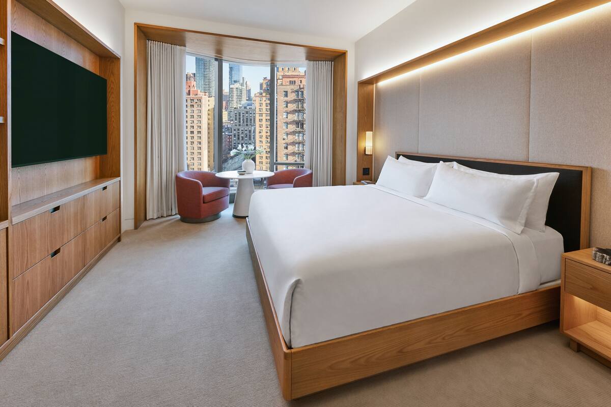 A hotel room with a large bed, a TV mounted on the wall, a small table with chairs, and a view of the city through large windows.