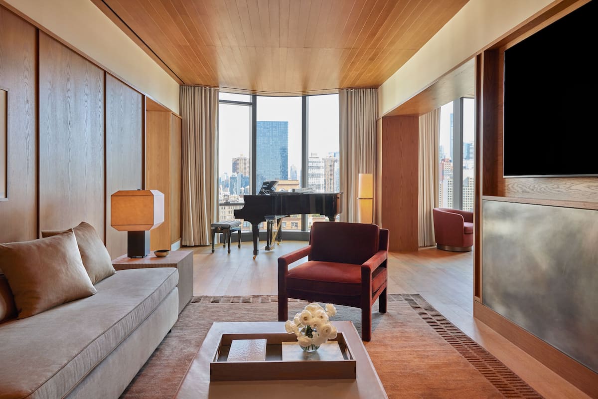 A modern hotel room with a large window, a piano, a red armchair, a sofa, and a flat-screen TV.