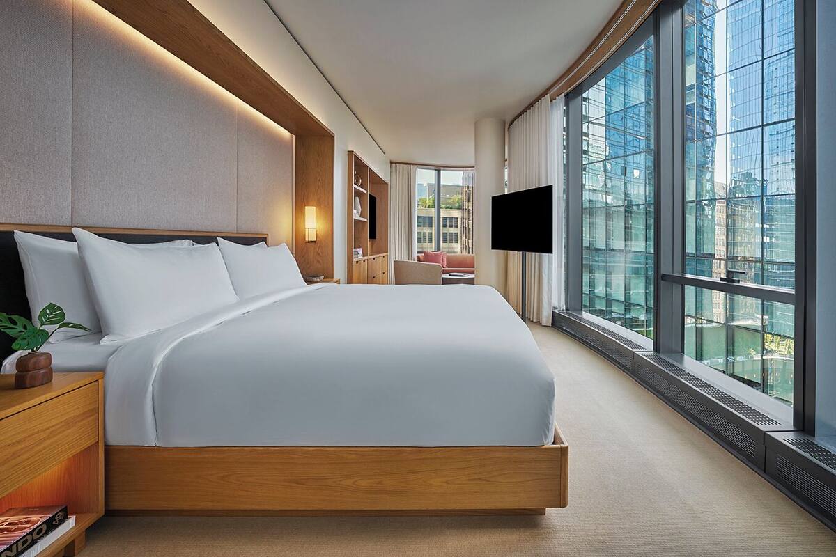 A modern hotel room with a large bed, a flat-screen TV, and a city view through large windows.