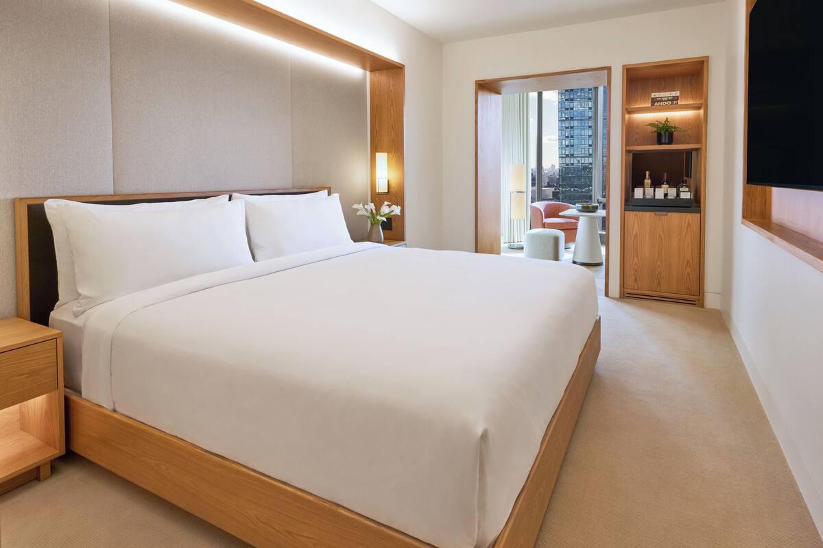 A hotel room with a large bed, a nightstand, a wooden wardrobe, a seating area with a chair, and a view of the city through a large window.