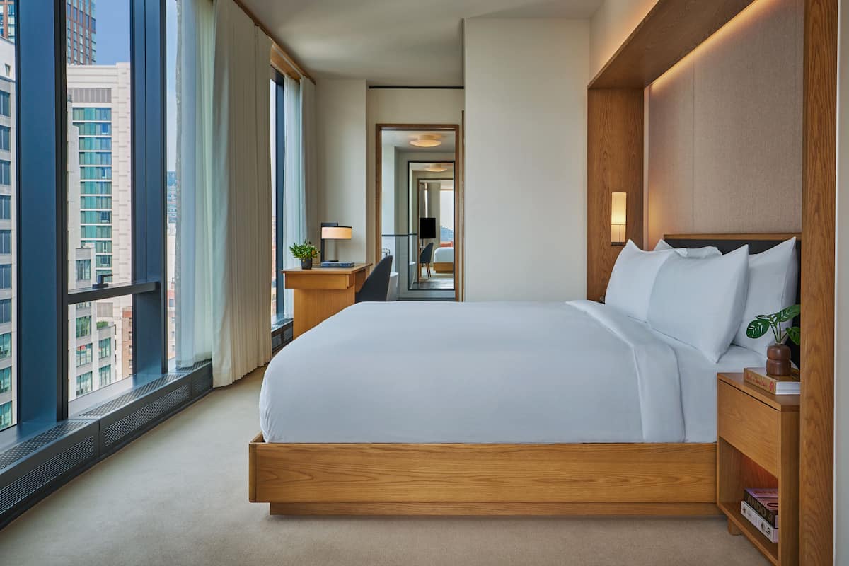 A modern hotel room with a large bed, a nightstand, a lamp, and a view of the city through large windows.