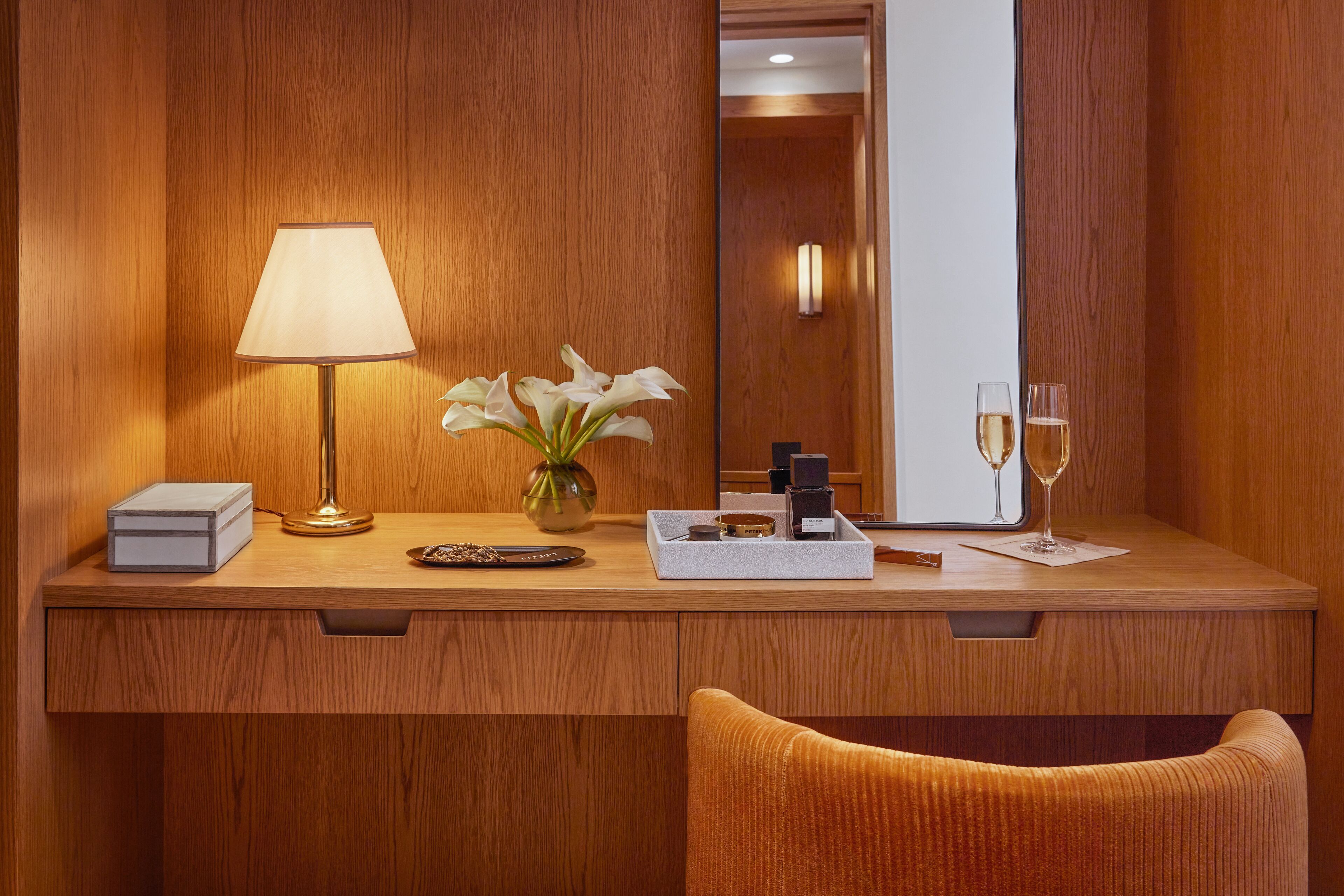 A wooden desk with a lamp, a vase with white flowers, a tray with a bottle and glasses, and a mirror reflecting a door.