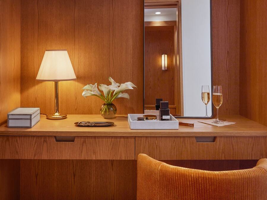 A wooden desk with a lamp, a vase with white flowers, a tray with a bottle and glasses, and a mirror reflecting a door.