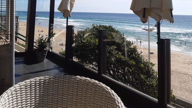Een balkon met uitzicht op het strand, met een witte parasol en een rieten stoel.