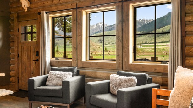 L’intérieur d’un chalet en rondins, avec deux fauteuils gris, une table en bois et une vue sur un paysage montagneux par de grandes fenêtres.