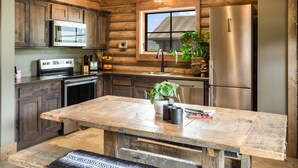Une cuisine rustique aux murs en bois, avec une grande table en bois, un réfrigérateur en acier inoxydable et une fenêtre donnant sur une maison.