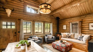 L’intérieur d’une cabane en rondins, avec une cheminée, des meubles en bois et de grandes fenêtres donnant sur le paysage extérieur.