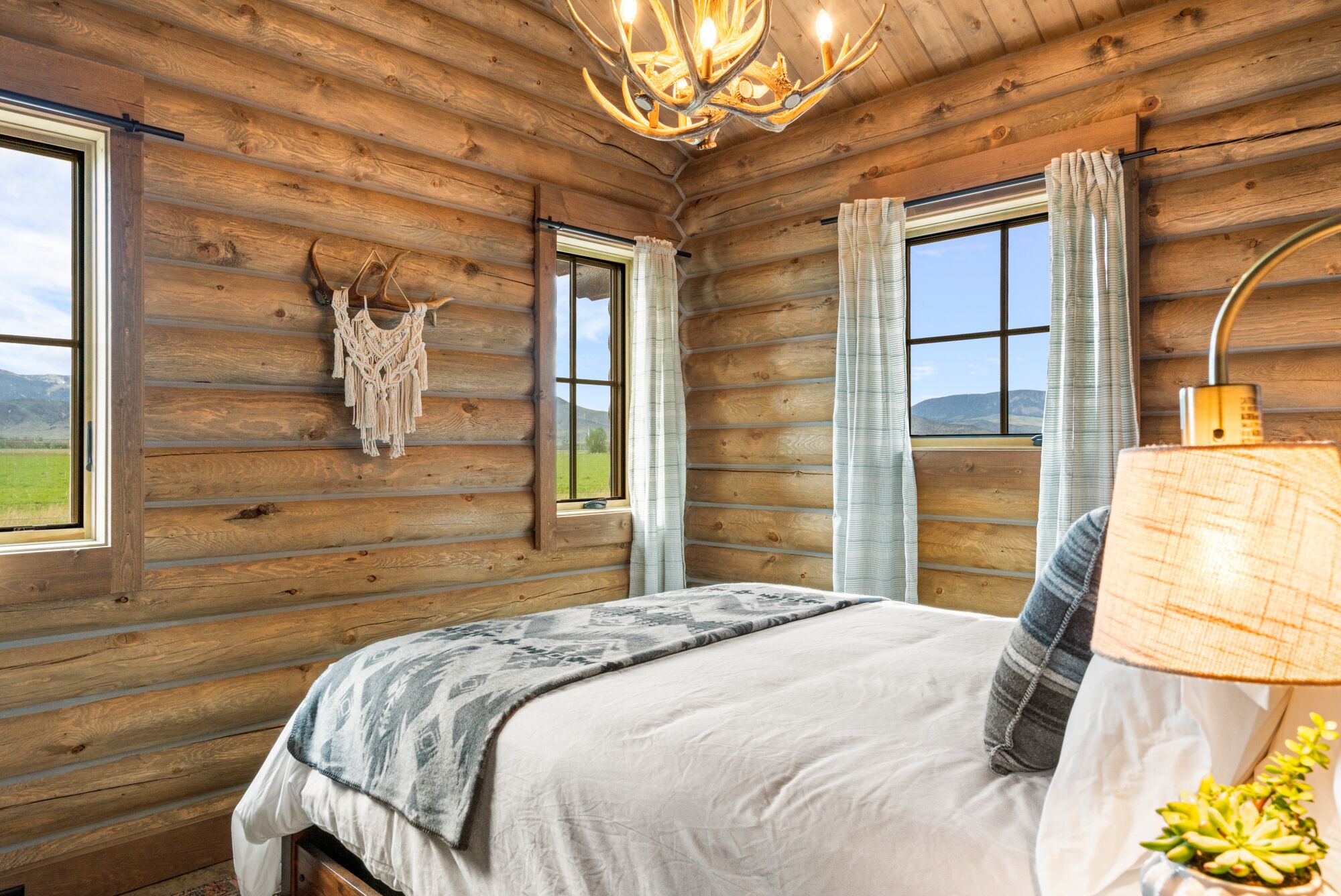 Une chambre à coucher aménagée dans un chalet en rondins, avec un lit, une décoration murale en macramé et une vue sur l’extérieur.