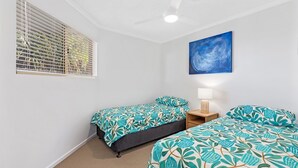 A bedroom with two beds, a ceiling fan, a window with blinds, a wall-mounted painting, and a bedside table with a lamp.