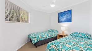 A bedroom with two beds, a ceiling fan, a window with blinds, a wall-mounted painting, and a bedside table with a lamp.