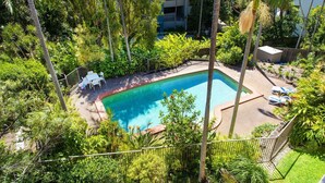 An aerial view of a swimming pool surrounded by lush greenery and palm trees, with white lounge chairs and a small building in the background.