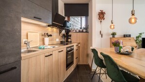 Ett modernt kök med träskåp, en inbyggd ugn, en kaffebryggare och en matplats med ett träbord och gröna stolar.