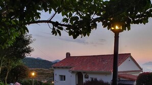Una casa con tetto in tegole rosse, circondata da un giardino con fiori di ortensia rosa, e un lampione in primo piano.