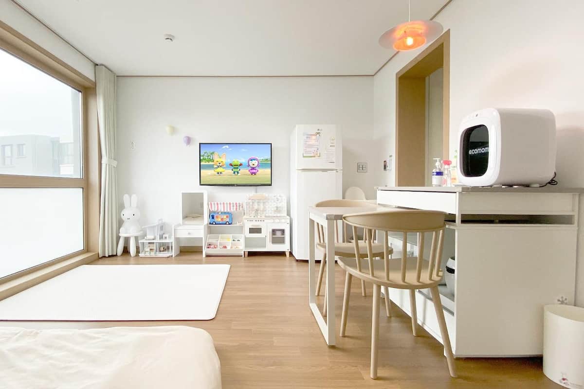 Ein modernes, helles Zimmer mit großem Fenster, einem Bett, einem Schreibtisch mit Computer, einem Stuhl und einem Fernseher.