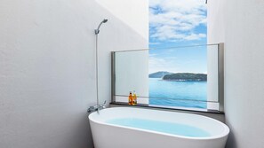 Een badkamer met een badkuip, een douche en een groot raam met uitzicht op de zee en een eiland in de verte.