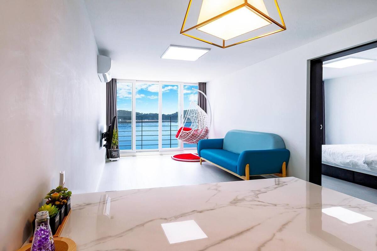 Habitación de hotel moderna con suelo de mármol, sofá azul, sillón colgante y vistas al mar a través de amplias ventanas.