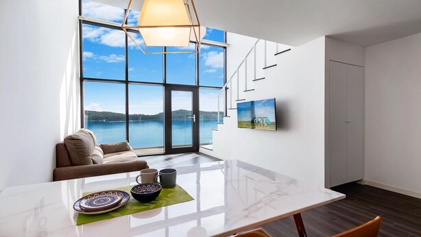 Ein modernes Wohnzimmer mit großem Fenster mit Seeblick, einer Couch, einem Esstisch mit dekorativen Tellern und einem Wand-Fernseher.