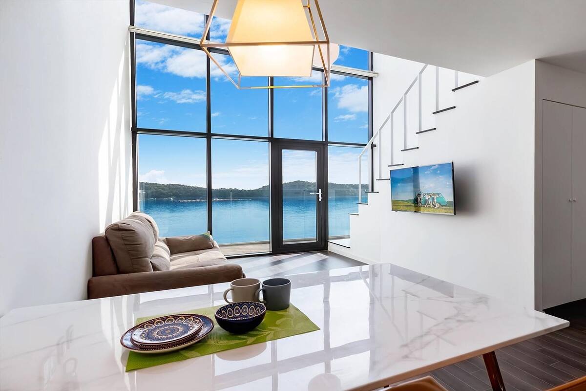 Una sala moderna con un ventanal grande con vistas a un lago, un sofá, una mesa de comedor con platos decorativos y un televisor montado.