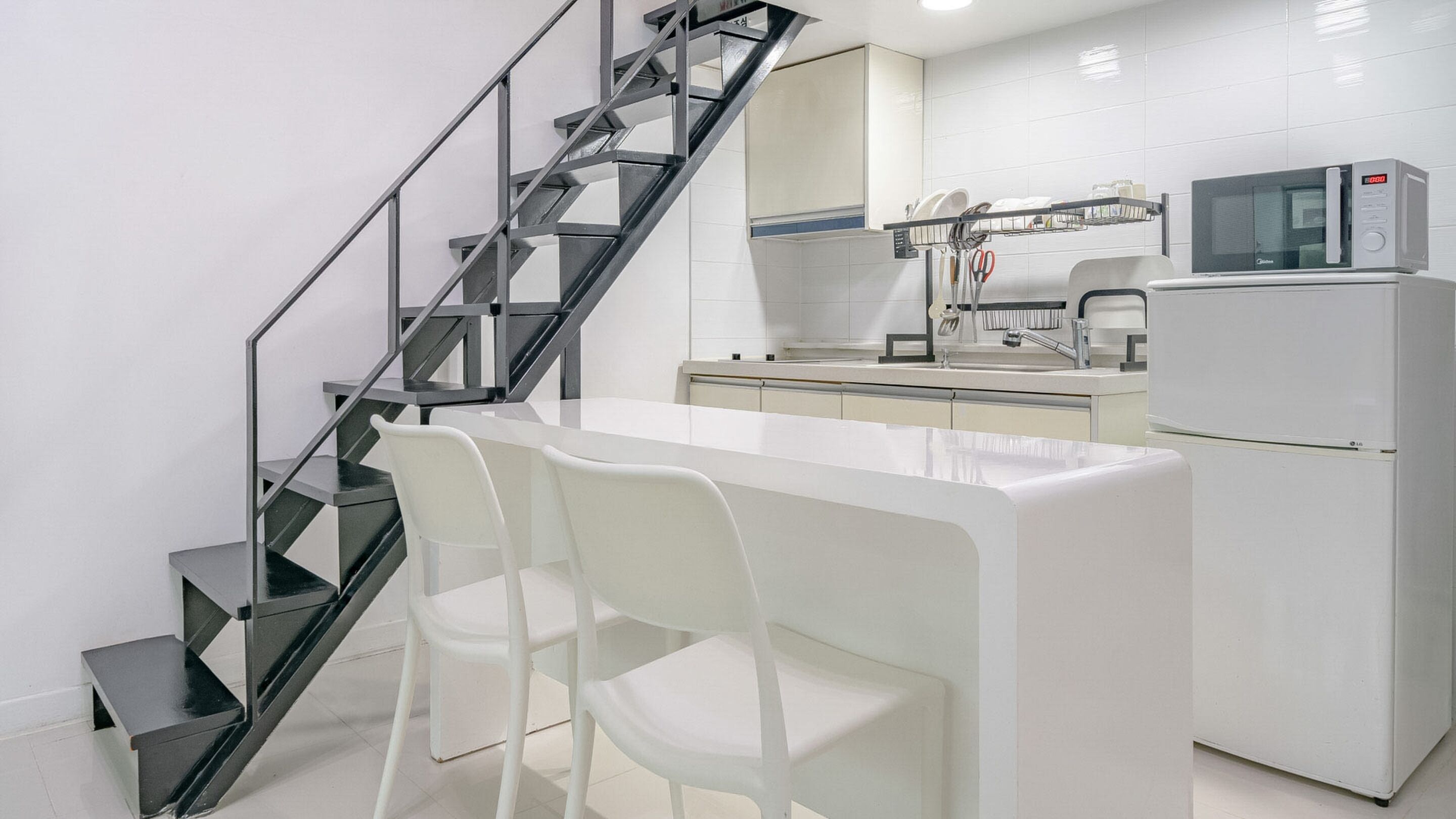 Una cucina moderna con un piano di lavoro bianco, un frigorifero in acciaio inossidabile, un forno a microonde e un forno integrato. Si vede una scala nera che conduce a una pedana con una sedia bianca.