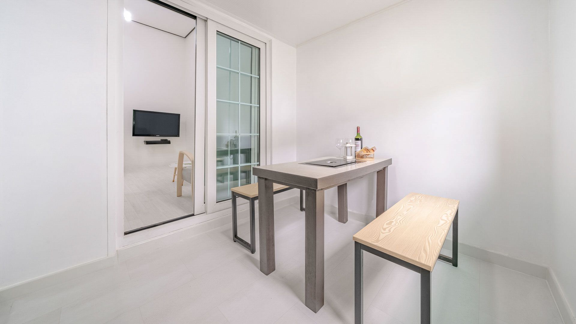 A modern, minimalist room with a glass door leading to a balcony, a small table with a bottle and glasses, and a television mounted on the wall.