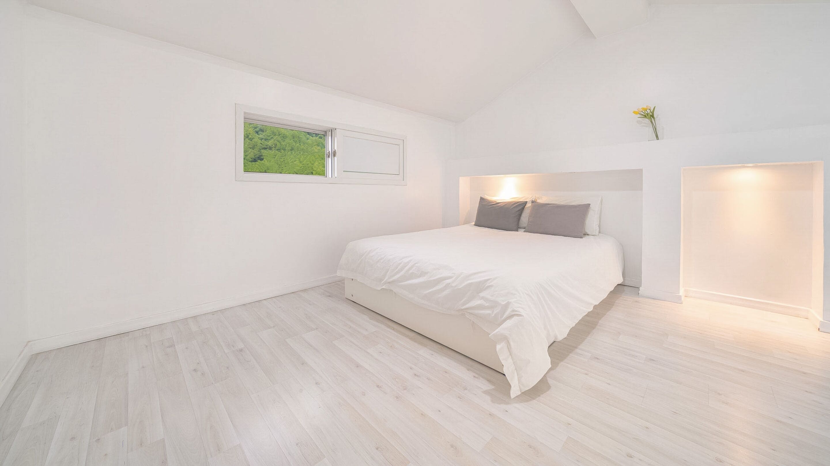 A minimalist bedroom with a large bed, a small window, and a nightstand with a vase of flowers.