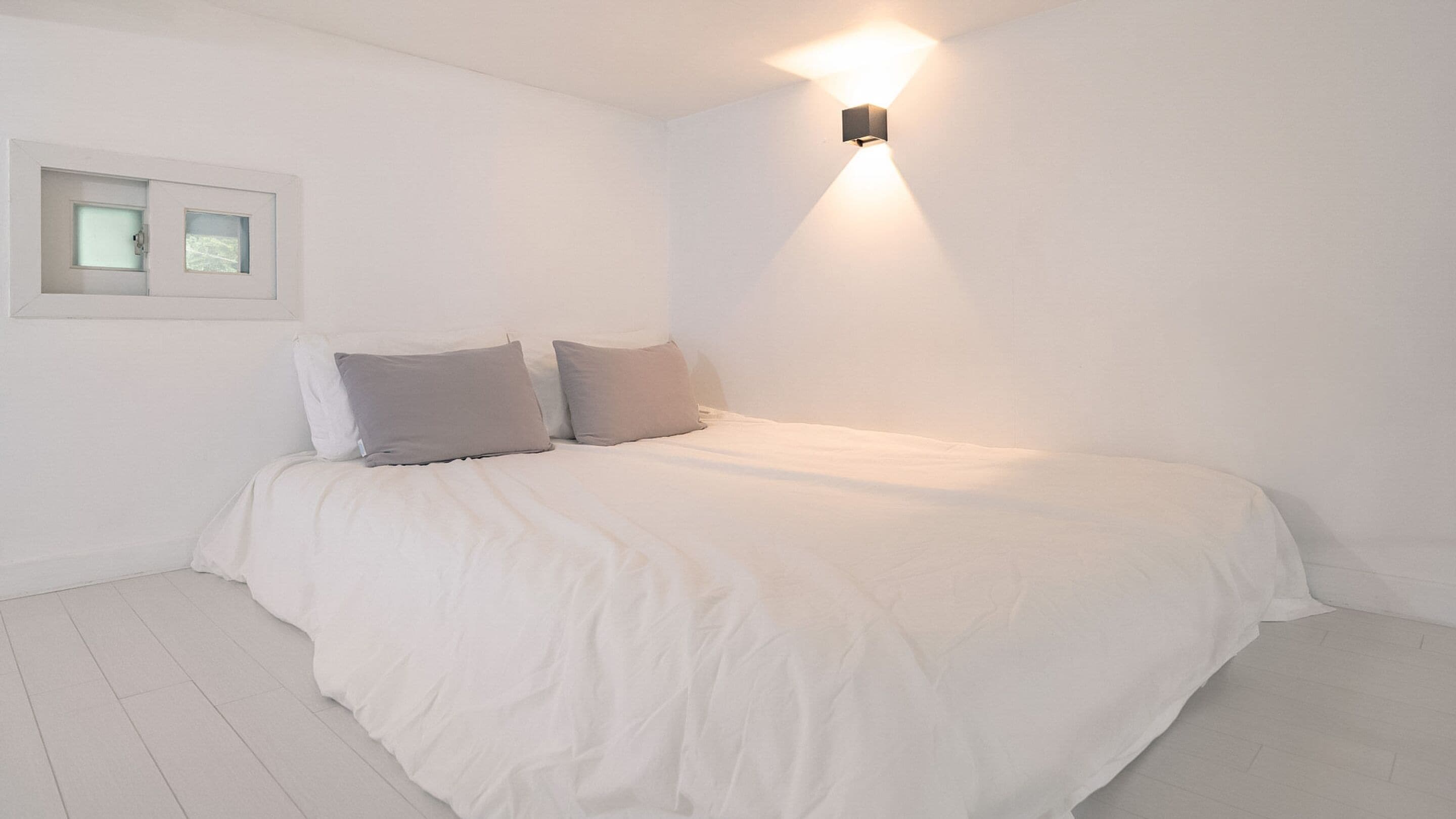 A minimalist bedroom with a large bed, white walls, and a small window.